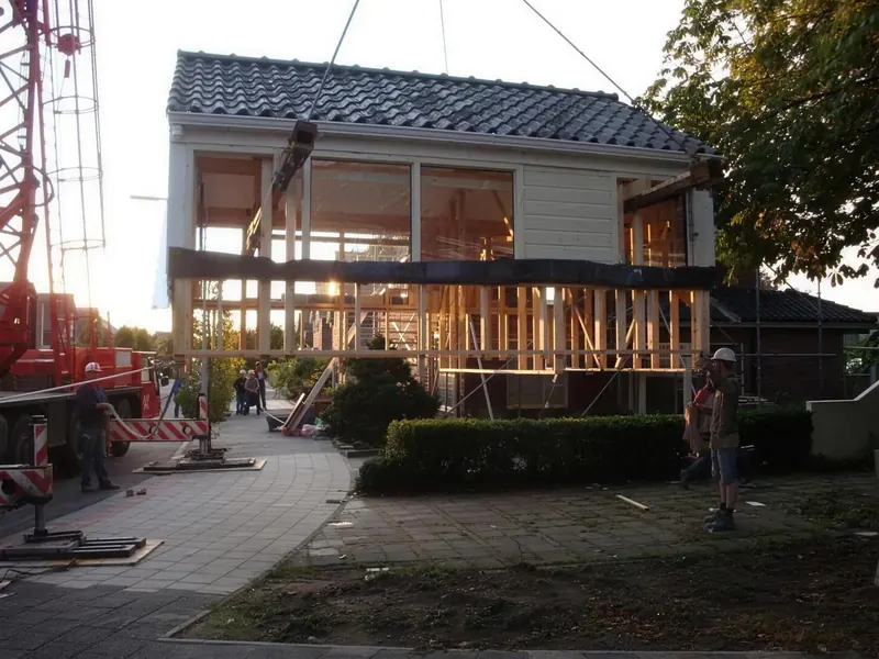 Dakopbouw met witte gevel en ramen hangt laag aan de kraan, medewerkers met helm ernaast