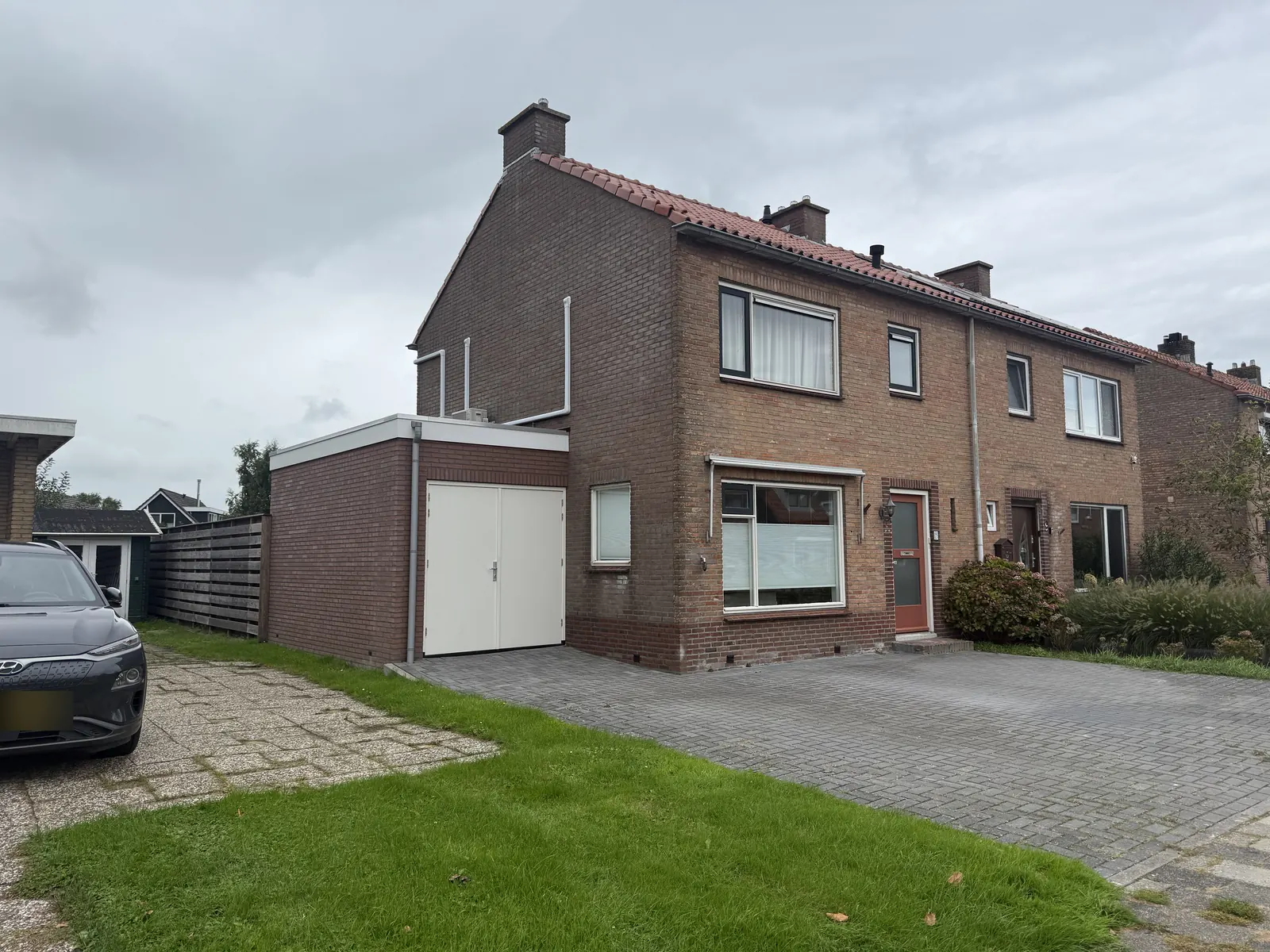 Garage van roodbruine baksteen met witte dubbele deuren naast een twee-onder-een-kapwoning in Spanbroek