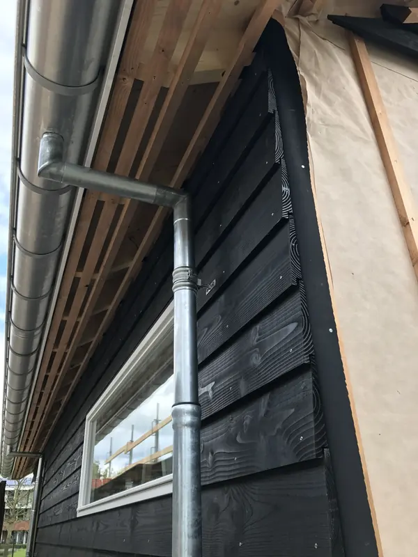 Zwarte houten gevelbekleding met zinken goot en regenpijp naast een wit kozijn, Hoogwoud