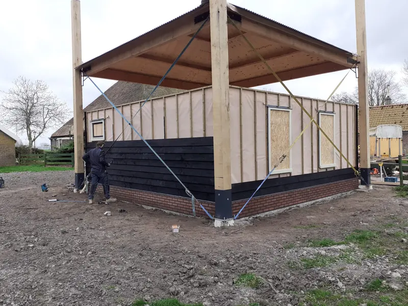 Kapschuur in Hoogwoud tijdens de bouw: zwarte gevelbekleding op een gemetselde plint, palen gespannen