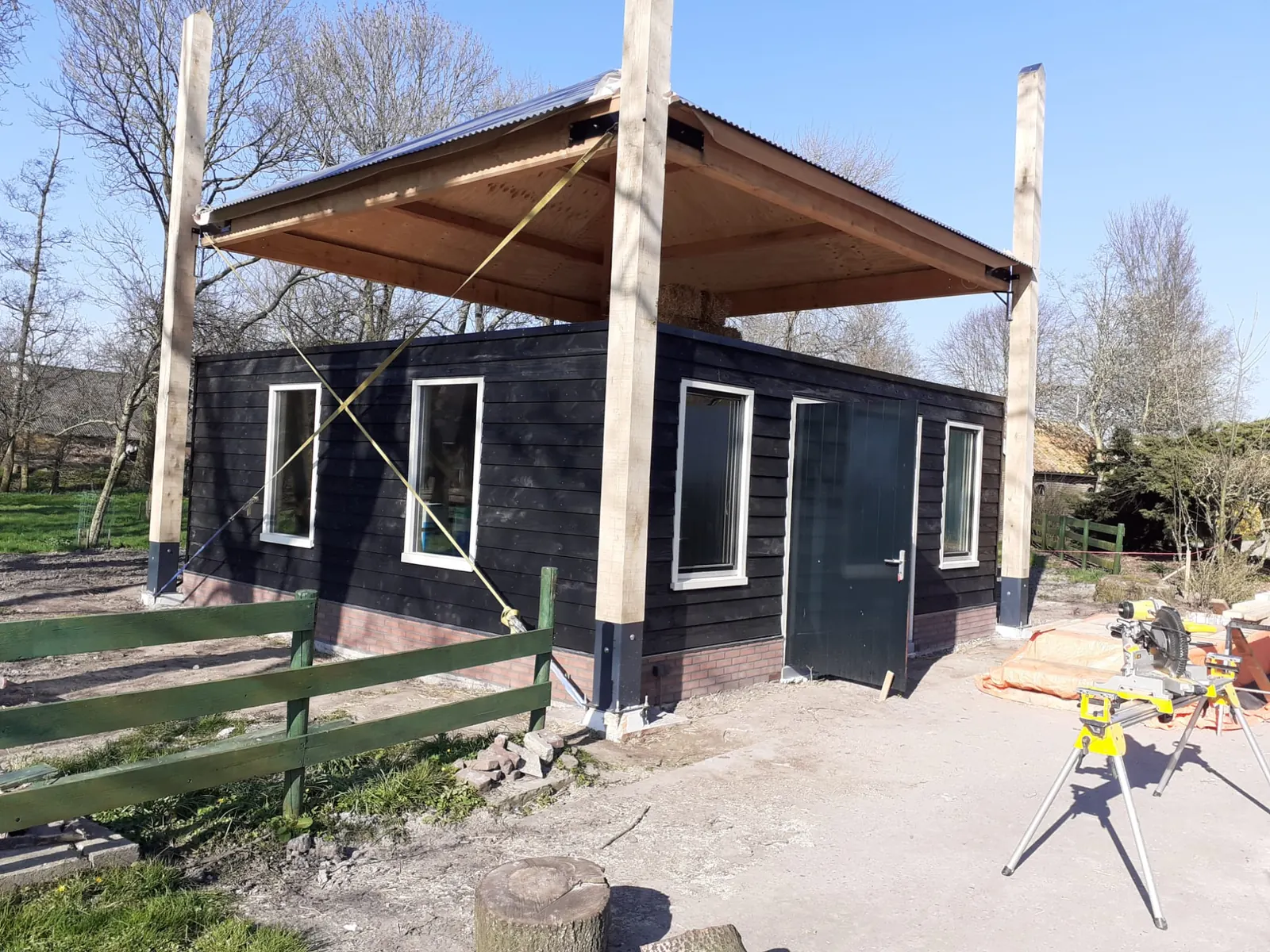 Kapschuur in Hoogwoud op vier houten palen met golfplaten kap boven een berging van zwarte delen