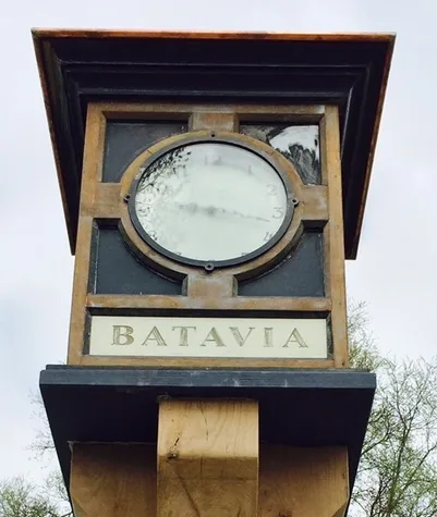 Klokkast met opschrift Batavia en koperkleurige kap op een houten paal