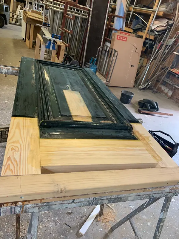 Groene paneeldeur met nieuwe houten onderregel en ingezet stuk in het paneel, op schragen in de werkplaats