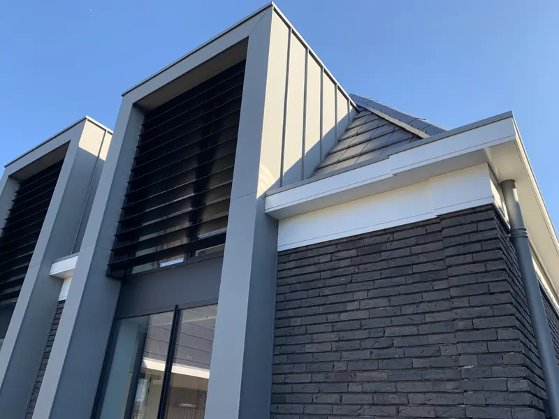 Twee hoge kaders van lichtgrijze plaat met zwarte lamellen naast donkere baksteen