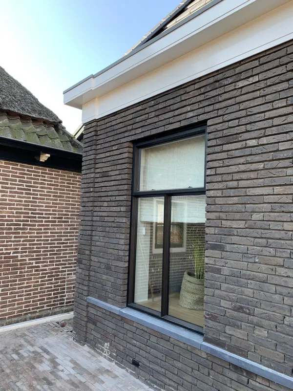 Zijgevel van donkergrijze baksteen met zwart kozijn en witte daklijst, woonhuis in Broek op Langedijk