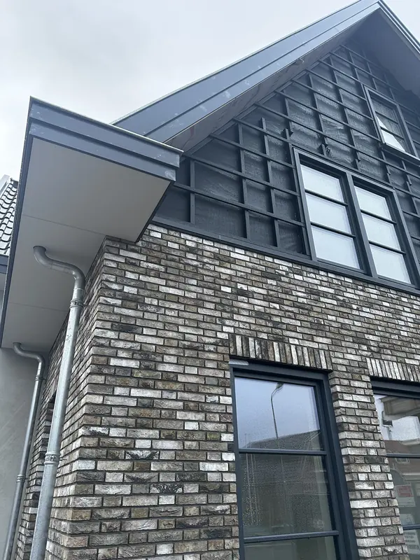Hoek van grijs gemêleerde baksteen met antraciet dakrand, zinken regenpijp en houten topgevel in Obdam